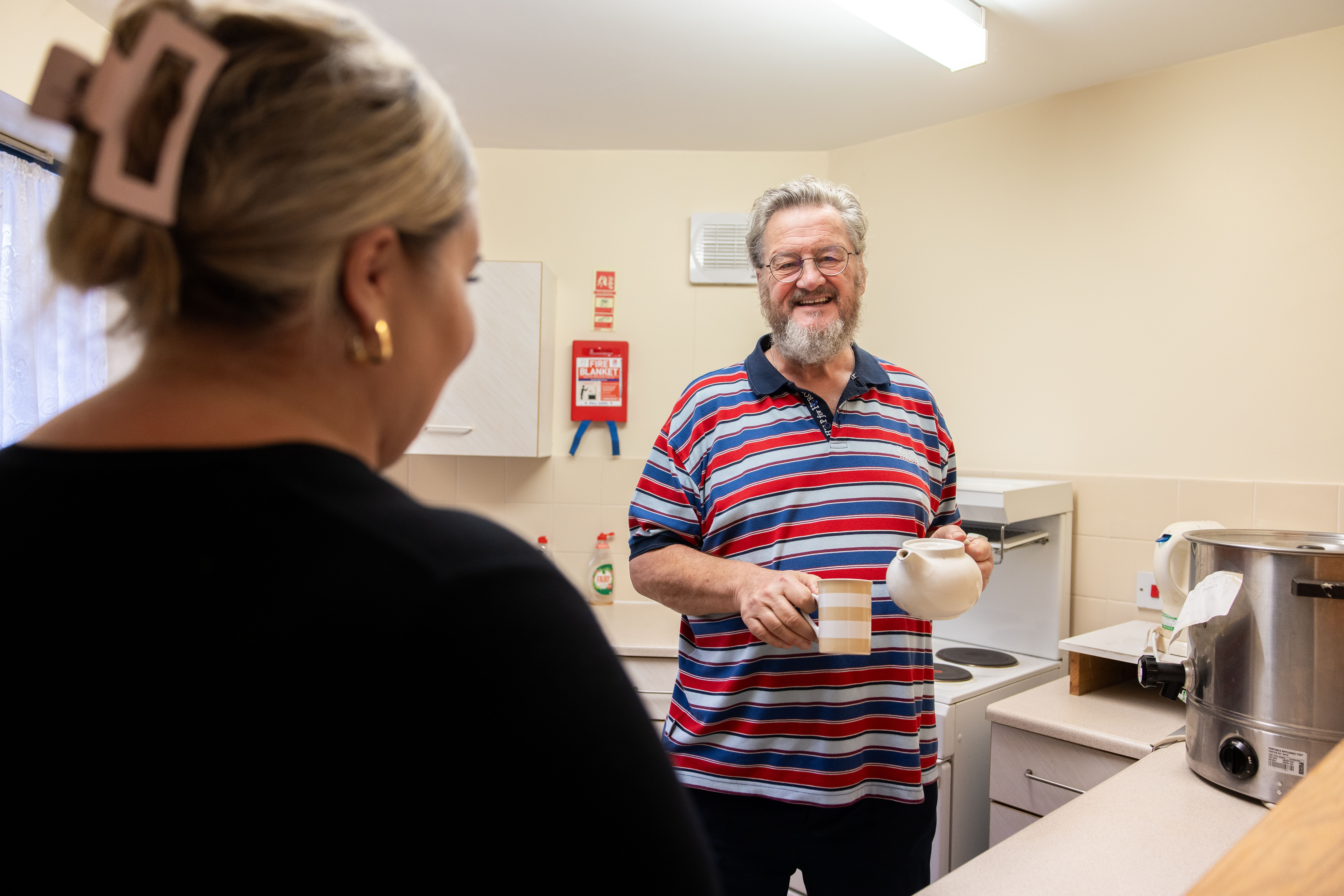 Man standing in a kitchen speaking to a member of staff holding a cuppa and a tea pot, looking at the camera and smiling