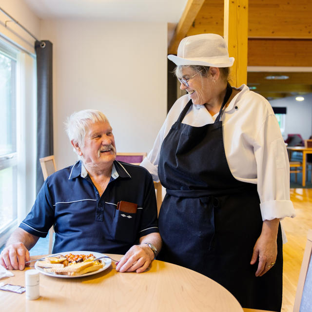 Older man sat having his breakfast, with a kitchen staff member talking to the man, with their arm around them, both are looking at each other and laughing and smiling. 