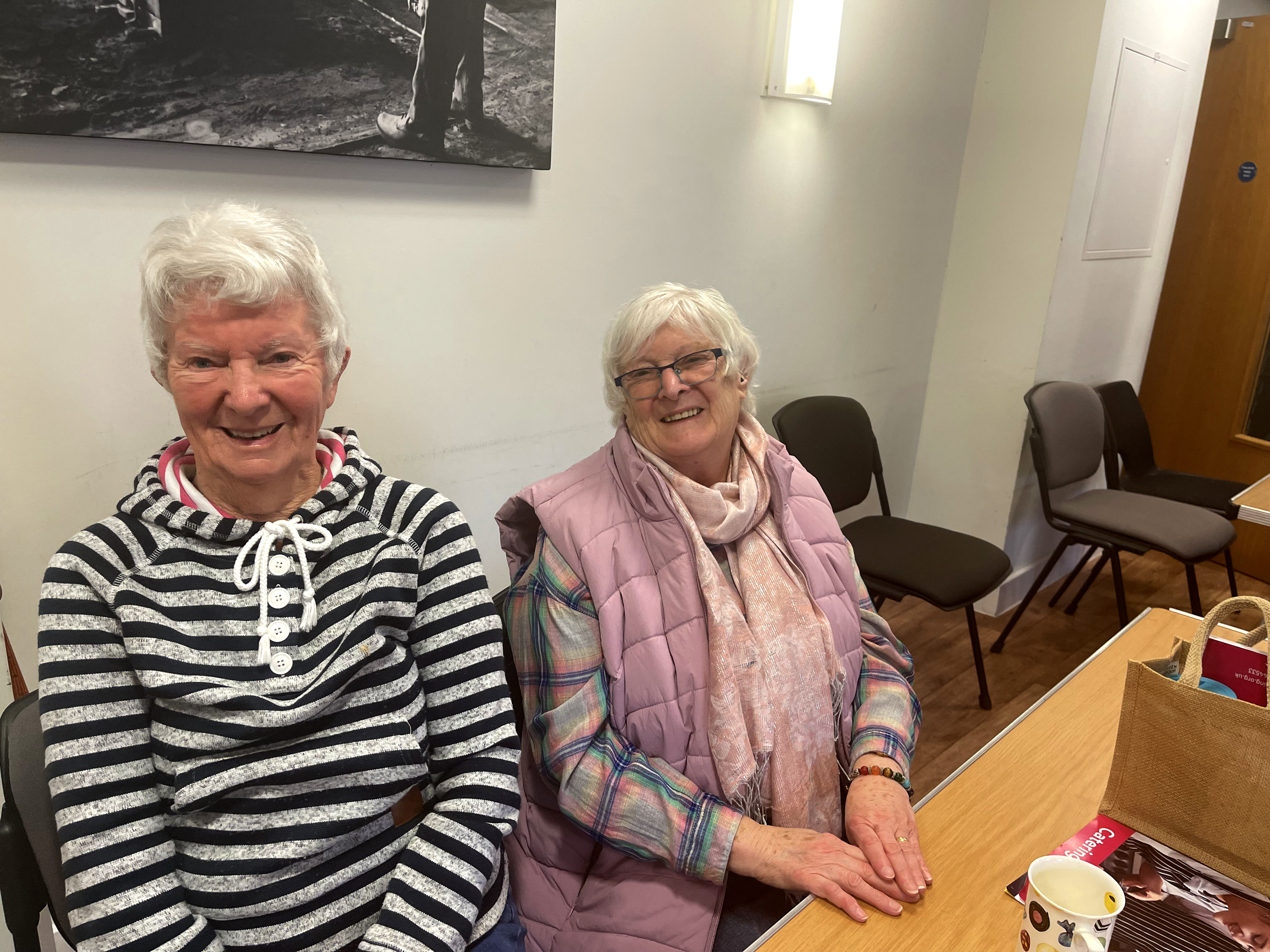Two older women sat next to each other smiling at the coffee morning