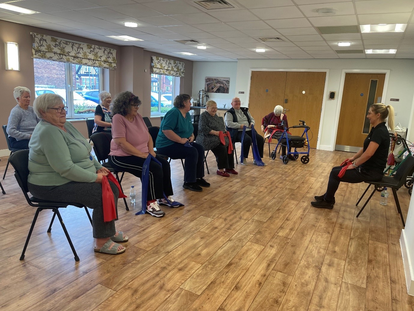 Residents in the community room at West End Village sitting down with resistance bands in their hands laughing and smiling