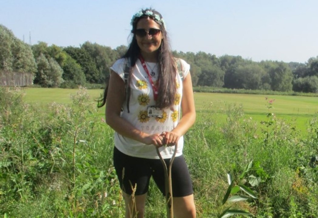 A woman standing in a field holding a shovel with a lanyard on smiling